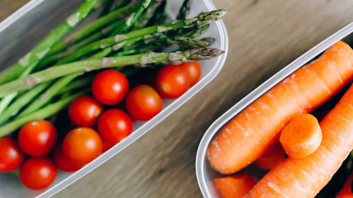 Close-up of fresh asparagus, cherry tomatoes, and carrots in plastic containers on a wooden table.